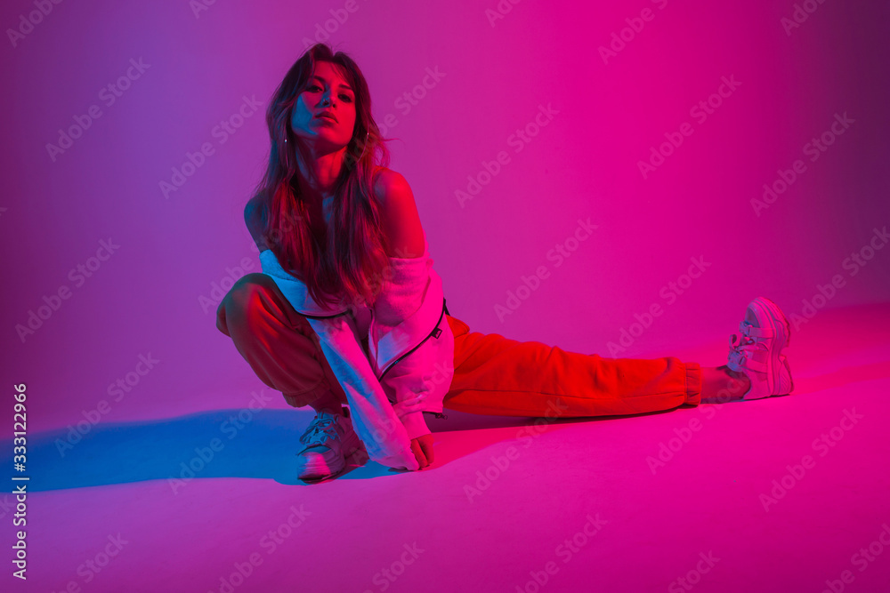 Wall mural Fashion pretty young woman in fashionable clothes is doing stretching indoors with neon bright red light. Modern girl posing in a studio with colorful color. Creative art concept. Art fashion. Style.