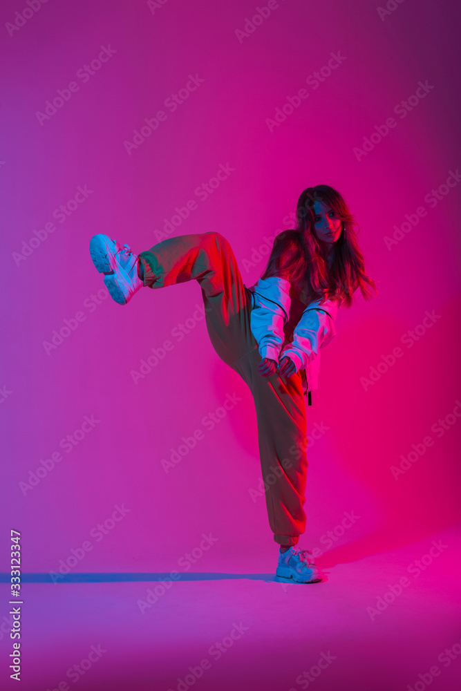 Poster Stylish young woman dancer with trendy sports youth clothes in fashion sneakers stands on one leg in the studio with a bright pink light. Modern girl dancing in the room with multi-colored neon color.