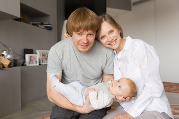 Cheerful positive new parents enjoying leisure time with baby daughter. Young couple holding six month child and looking at camera. Parenthood concept