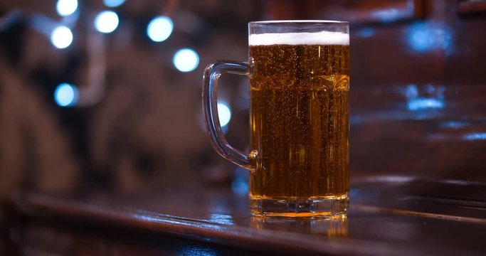 Close up of one half liter glass mug full of beer standing on a brown varnished wooden bar counter in a dark pub with blinking lights in the background. With free space for writing 4K