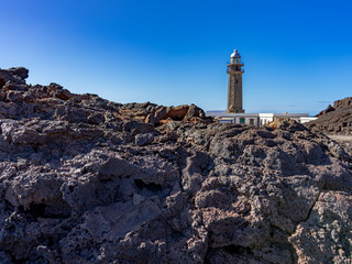 In the foreground volcanic rock, a very common landscape on the coast of the island of El Hierro.  In the background, out of focus is the La Orchilla Lighthouse.