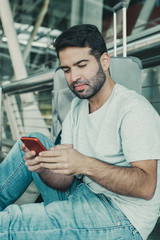 Smiling young guy sitting on floor and looking at phone. Cheerful handsome bearded Hispanic man using smartphone at airport. Travel concept