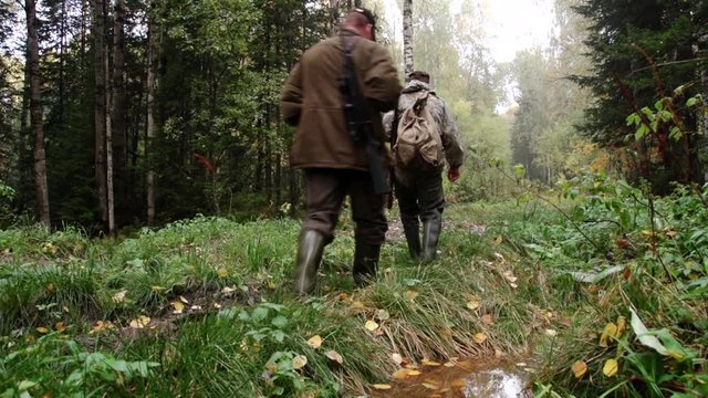 Two hunters go through dense autumn forest, hunting grounds, wet land, puddles, lot of autumn foliage, rifle with an optical sight on shoulder, dirt and cold, carry supplies in large tourist backpack