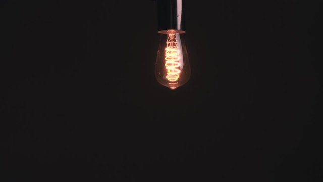 Dark grey gradient background, vintage LED light bulb turns on, off, flickers, glows, hanging off the ceiling in the dark  room.