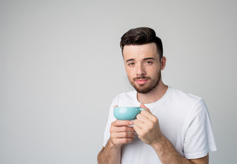 Young man isolated over background. Casual guy in white shirt hold blue cup in hands with tea or coffee inside. Loook straight on camera and pose. Ready to drink warm or hot drink.