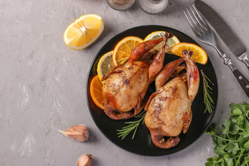 Baked quails with lemon and orange served on a dark plate on a gray background, Copy space