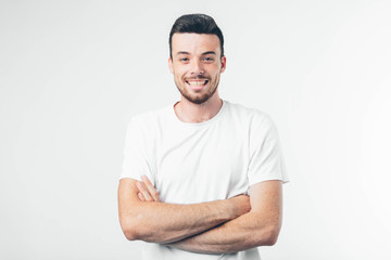 Fototapeta premium Young brunette man with beard dressed in white T-shirt. Guy confident and wide smiling. Arms crossed at chest level. Isolated over light background.