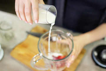 A female hand pours milk into a blender to prepare a tasty and healthy smoothie of strawberries and...