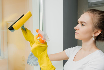 Cute adult girl washes a window with a scraper. Spring cleaning. An employee of a cleaning company wipes plastic windows in the house