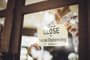 Store owner turning close sign broad through the door glass for social distancing prevent corona...