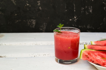 Slices of ripe watermelon on a plate and red watermelon juice in a glass on a wooden white background, and a black wall - side view. There is a place for text.