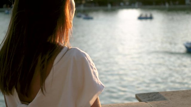 Rear view of attractive happy girl looking at boats sailing across a duck pond in urban park at sunset in the background. Madrid. Spain. 4K