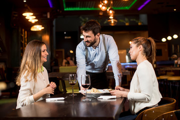 Smiling young female friends at a restaurant with waiter serving dinner