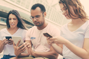 Group of young people using devices on street. Calm friends sitting with smartphones and tablet. Technology concept