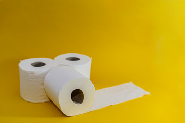 A stack of three white disposable toilet rolls on a yellow background. One roll lies untwisted. Soft fresh cotton. Essential goods. Purchasing power. Copy space.