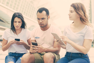 Young people sitting on street with digital devices. Front view of focused friends using tablet and smartphone. Technology concept