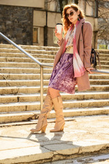 Photo of a fashionable glamorous Beautiful woman with long hair with a cup of coffee in the spring city in the sun. A model stands outdoors on the background of the steps of the building