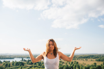 Young woman practices yoga outside. Calm blonde girl with closed eyes. She dressed in white T-shirt. Her palms turned to sky. Portrait. Trees river and sky on background.