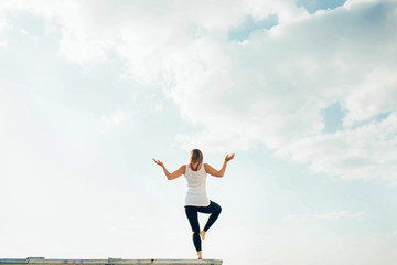 Young woman practices yoga outside. Blonde girl turned back standing on one leg, second raised bent at knee. Hands on sides bent at elbows and turned up. Sky and clouds on background.