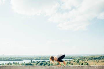 Young woman practices yoga outside. Blonde girl do crane pose or bakasana on parapet with beautiful view. She dressed in black leggings and white T-shirt. Trees river and sky on background.