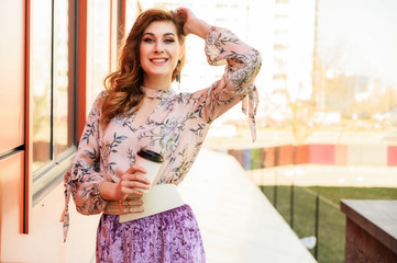 Photo in bright weather of a fashionable glamorous beautiful woman with long hair against the background of the spring city in the sun. Model is standing outdoors with a cup of coffee in her hands.