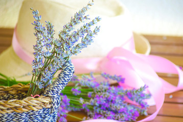 Light straw hat on a wooden brown table. Lilac-pink ribbon on the hat of a woman's hat. A bouquet of dry lavender in a rustic wicker straw basket.