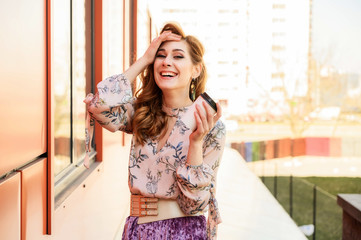 Photo in bright weather of a fashionable glamorous beautiful woman with long hair against the background of the spring city in the sun. Model is standing outdoors with a cup of coffee in her hands.