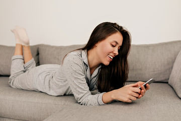 Woman in pajamas lying on sofa with smartphone and scrolling. European girl resting at home