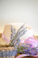 Light straw hat on wooden brown table. Lilac-pink ribbon on hat of woman's hat. A bouquet of dry lavender in rustic wicker straw basket.