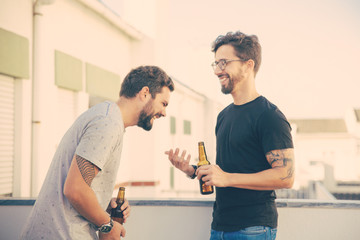 Two male friends drinking beer and sharing funny news on outdoor terrace. Two young men in casual...