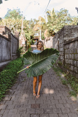 Beautiful and young slender girl poses as a model with a leaf on the island of Bali. The culture and nature of a tropical island is conveyed. Emotions and happiness