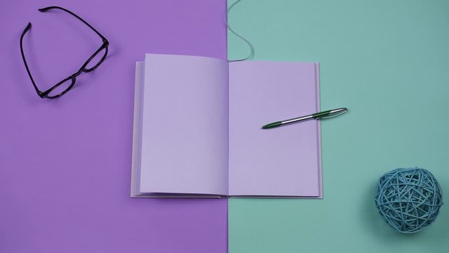 Video of a book's page turning stop motion on colored background with different office stuff. Blank template for words inside an open purple notebook. Writing and creative concept