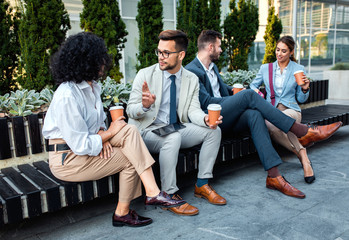 Fototapeta premium Group of coworkers having a coffee brake together, siting outside in front of office buildings and talking.