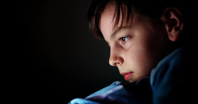 Video of a child holding a mobile phone and watching a movie, at night, with his face illuminated.