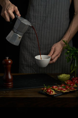 Chef man hands holds coffee pot and pouring coffee into a white cup. Dark style photo.