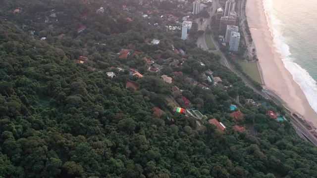 Aerial View Of Parachuter Gliding Over Lush Green Forest Toward Beach At Sunrise