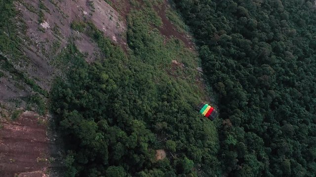 Aerial View Of Parachuter Gliding Down Over Large Rock Face And Lush Green Forest