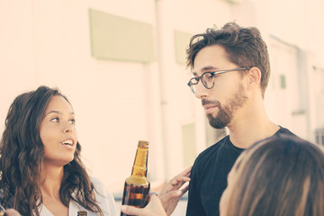 Group of friends discussing news over bottle of beer on outdoor terrace. Young men and women in casual meeting outside. Friendly hangout concept