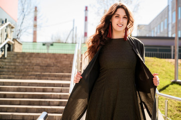 Obraz premium Photo of a pretty fashionable brunette woman with long beautiful hair with a smile in a suit on the background of the steps of the building in sunny weather