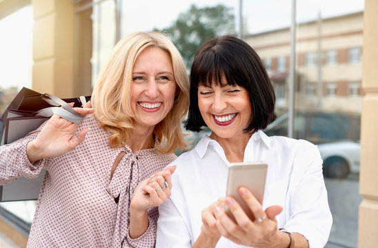 Two mature women looking at the phone and having fun while doing shopping