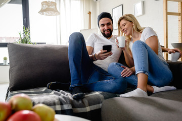 Obraz premium Young hipster couple sitting on sofa at home talking, drinking coffee, looking at smartphone
