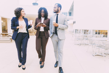 Business colleagues chatting while walking through office hallway. Business man and women using mobile phone, drinking coffee during work break. Corporate discussion concept