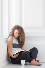 Vertical picture of young woman with curly hair sitting on floor in white woom alone. Look down at cup. Sit with crossed legs. Portrait.