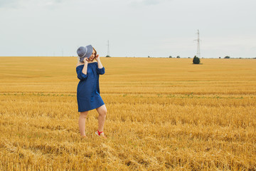 Young beautiful blonde girl in the middle of a wheat field. Posing looking at the camera. Summer landscape, good weather. Blue cotton dress, eco style. Beautiful striped hat.