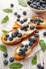 Sweet toasts with creamy vanilla cheese, honey and fresh blueberries on a white wooden table, close up. Delicious breakfast or dessert