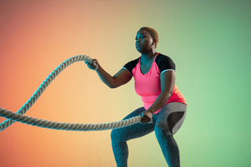 Young african-american plus size female model's training on gradient background in neon light....