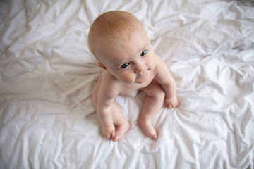 Adorable 6 month old baby sat on a bed looking up at the camera