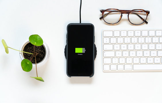 Concept of wireless charging, top view of phone charging next to the keyboard, cute plant and glasses