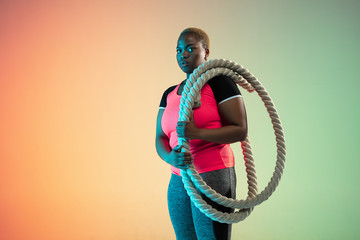 Young african-american plus size female model's training on gradient background in neon light....