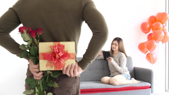 Over exposure scene of young adult asian girl in informal dress pay attention at notepad sit on sofa was surprised by boyfriend with gift box hide behind his back, newlywed sweet time, new pair family